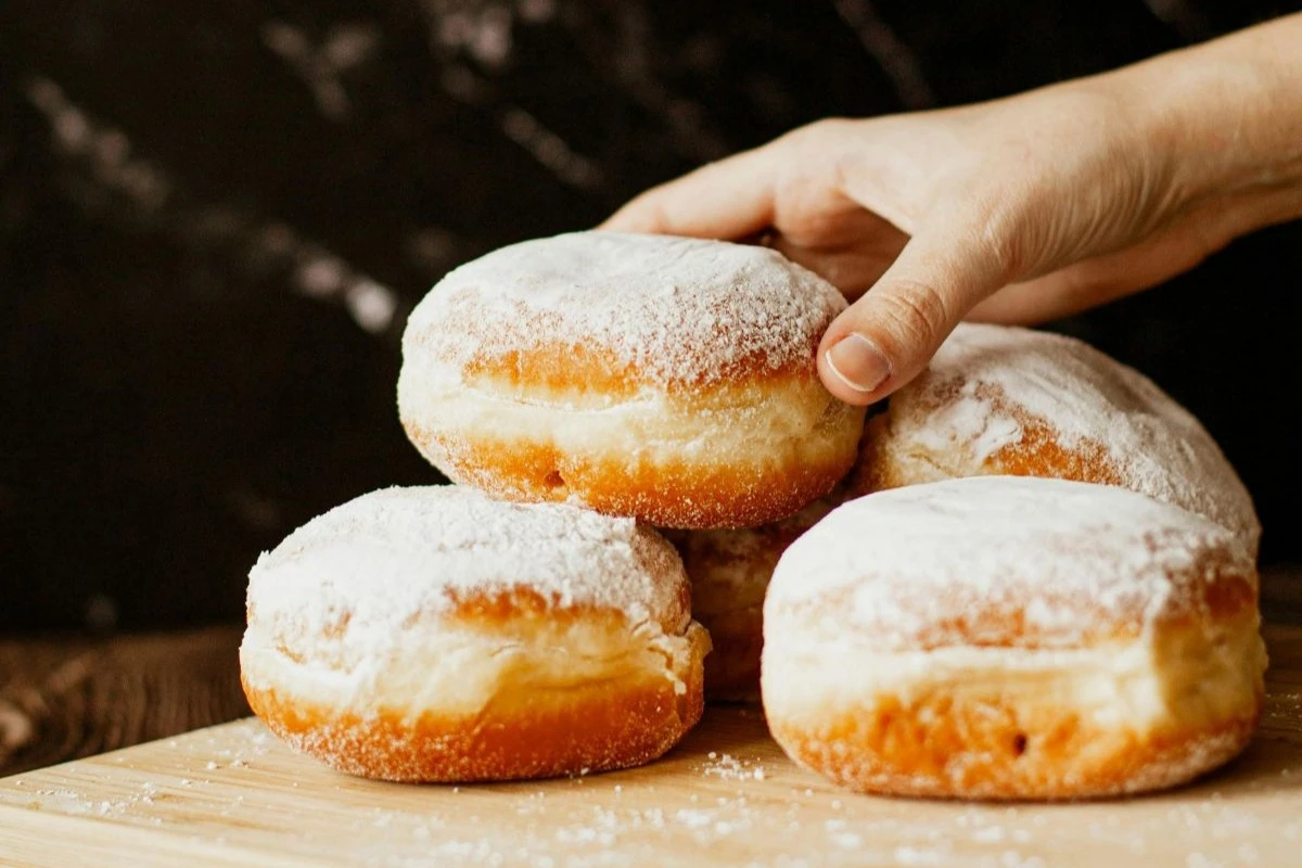 Vier übereinander gestapelte Krapfen, die mit Zucker bestreut sind, und eine Hand, die nach dem obersten Krapfen greift