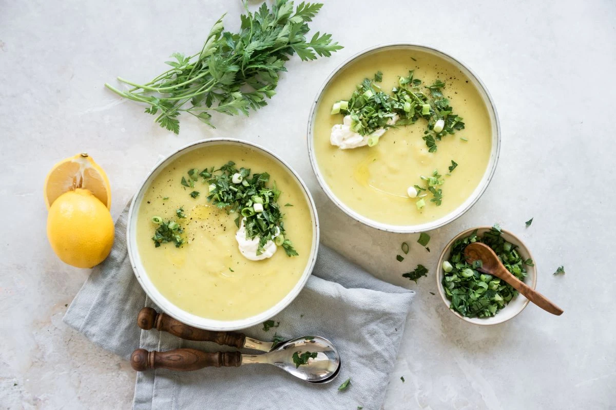 zwei Suppenschalen mit Erdäpfel-Lauch-Suppe, daneben diverse Kräuter und Frühlingszwiebeln und eine aufgeschnittene Zitrone